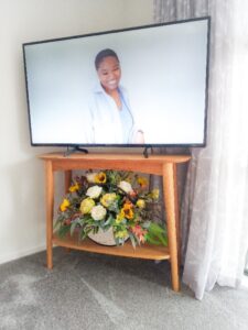 A hall table in a corner with Television on top and flower arrangement on the shelf underneath 