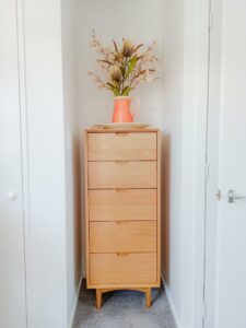 A slim oak of drawers fitting into a small nook. A peach ceramic vase of flowers sits on top of the drawers. 
